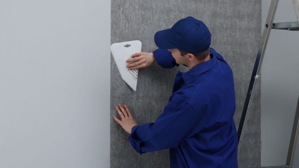 Person in blue work uniform smoothing wallpaper on a wall using a plastic scraper, with a ladder visible nearby.